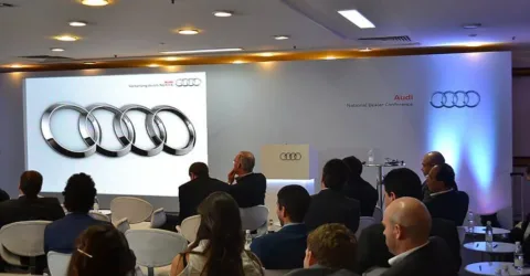 Painel de LED exibindo a marca Audi em conferência corporativa com plateia